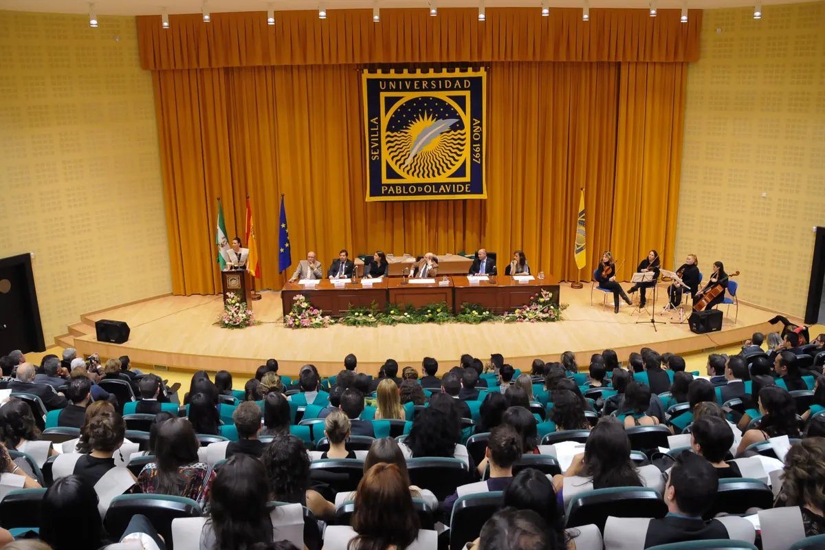 Acto académico en un salón de actos de la Universidad Pablo de Olavide, con el emblema de la universidad al fondo